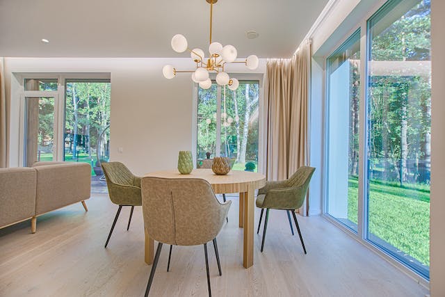 A modern dining area with a round wooden table, four upholstered chairs, and large windows offering a view of a green outdoor space. A stylish chandelier hangs above the table.