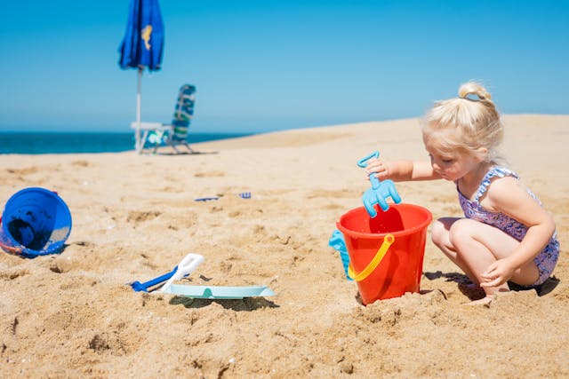 a-young-child-plays-in-the-sand-at-the-beach-scooping-sand-into-a-red-bucket-with-a-blue-toy-shovel-beach-toys-and-chairs-are-visible-in-the-background-under-a-clear-blue-sky